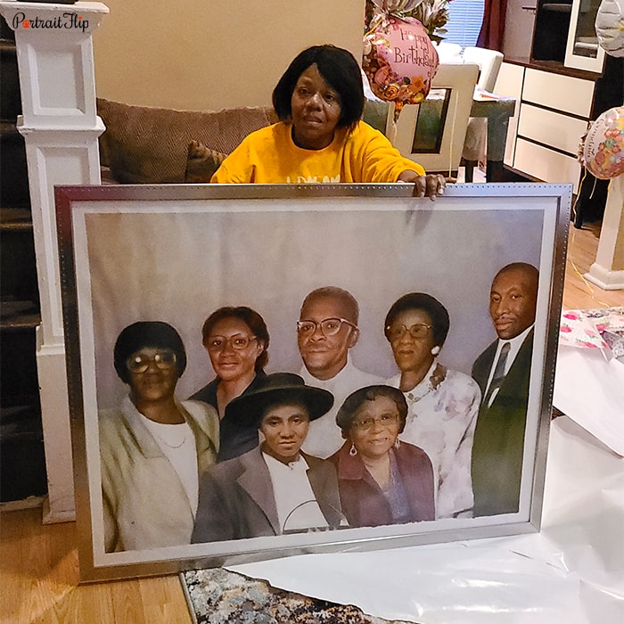 Picture of a woman holding family portraits that include two women in the left corner, two women in the front, a man in the right corner and an old man and woman beside him