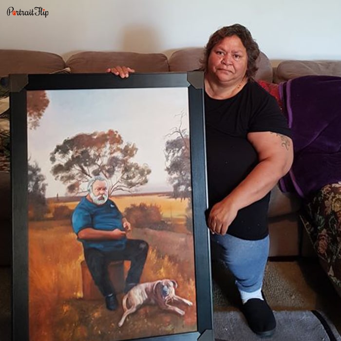 Picture of a woman holding mother’s day paintings, which include a man sitting in a forest view with his dog beside her legs